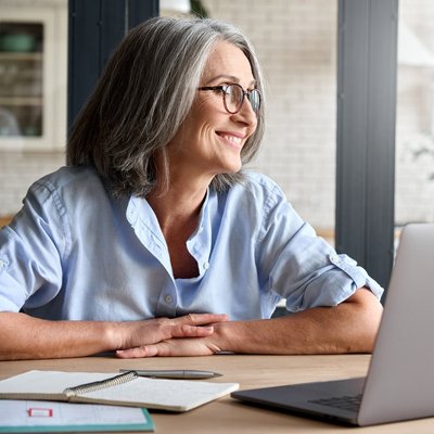 Frau am Laptop lächelt und schaut aus dem Fenster. Eine ältere Frau mit Brille sitzt an einem Tisch mit einem Laptop.