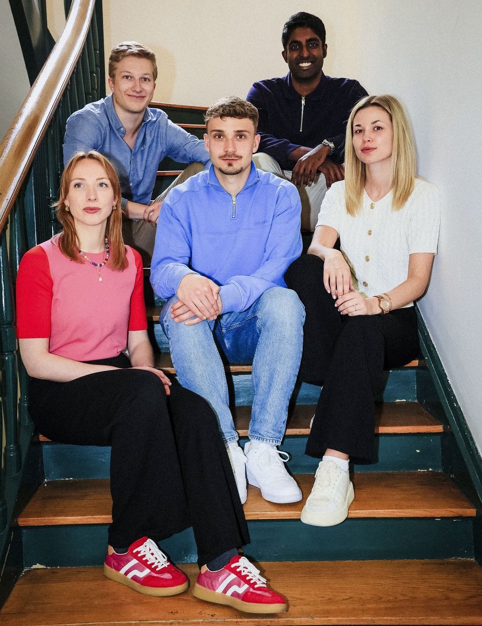 Fünf junge Erwachsene, zwei Frauen und drei Männer, sitzen in Freizeitkleidung auf einer hölzernen Treppe..