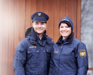 Eine lachende Frau und ein lachender Mann in Justizuniform stehen vor einem Tor.