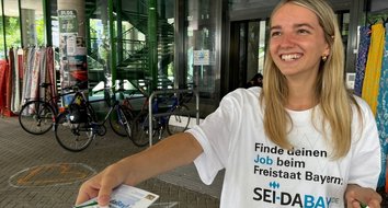 Promoterin am 9. Juli 2024 an der Uni Augsburg Auf dem Bild ist eine Promoterin zu sehen, die ein weißes T-Shirt mit dem "Sei-Dabay"-Logo trägt und einen Flyer an eine andere Person gibt.