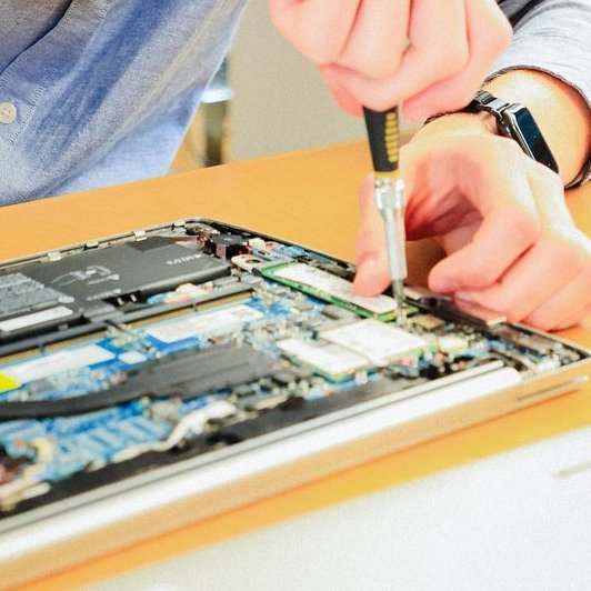 Ein junger Mann repariert lächelnd einen geöffneten Laptop an einem Schreibtisch.