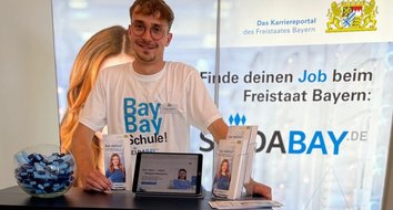 Ein junger Mann mit Brille und Namensschild steht lächelnd hinter einem Informationsstand für das Karriereportal des Freistaates Bayern „Sei-daBay.de“. Er trägt ein weißes T-Shirt mit dem Slogan „BayBay Schule!“. Auf dem Tresen befinden sich ein Tablet mit Informationsseite, mehrere aufgestellte Flyer und eine große Glasschale mit blauen und schwarzen Giveaways. Im Hintergrund steht ein beleuchteter Aufsteller mit dem bayerischen Staatswappen und dem Text „Finde deinen Job beim Freistaat Bayern“.