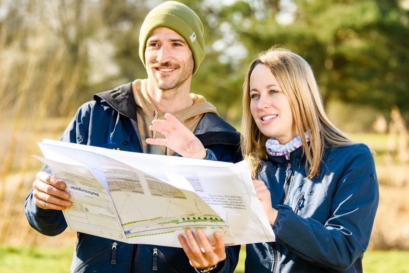 © Bayerisches Staatsministerium für Wohnen, Bau und Verkehr - Bildrechte: Uwe Niklas Eine lachende Frau und Mann schauen auf einen Plan.