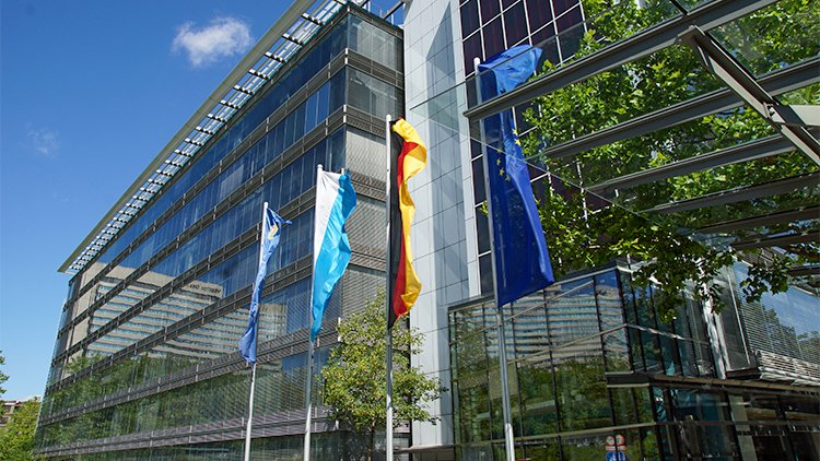 © Bayerischen StMUV Gebäude mit Glasfassade mit einer Deutschland-, EU- und Bayernflagge im Vordergrund.